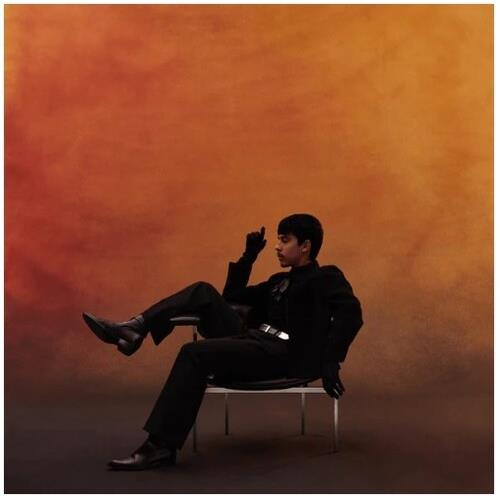 Young man in black suit seated on a white chair against an orange background