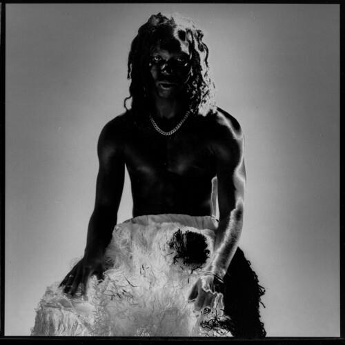 Black and white portrait of a shirtless man with dreadlocks wearing a pearl necklace and feathered s