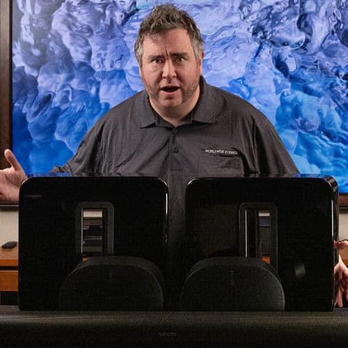 Man in gray shirt presenting two black speakers with visible drivers