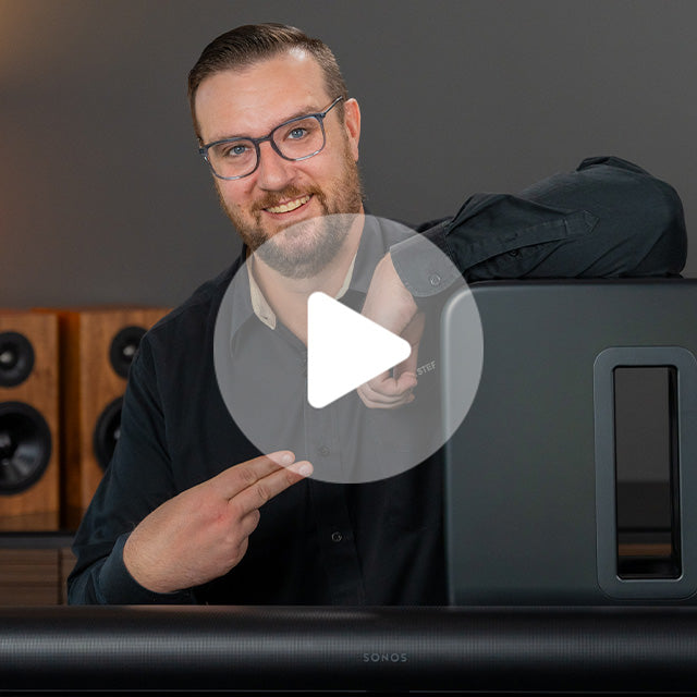 Man with glasses and beard smiling next to a black Sonos speaker