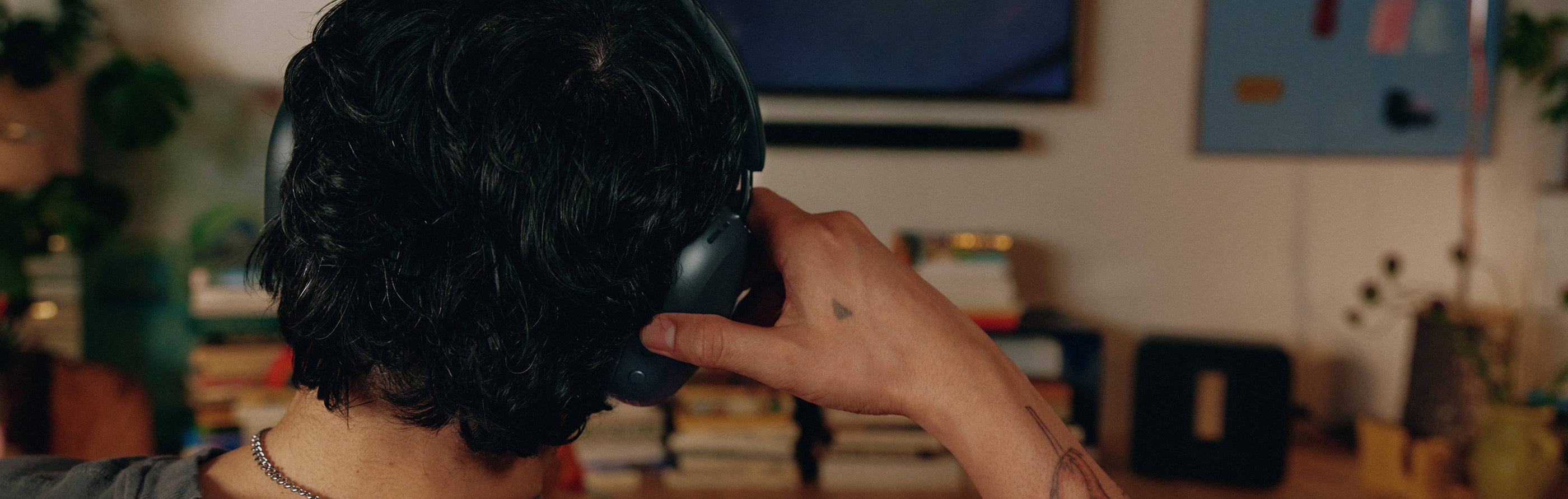 Man adjusting the Sonos Ace Ultra headphones from behind while a soundbar and TV are visible on the wall