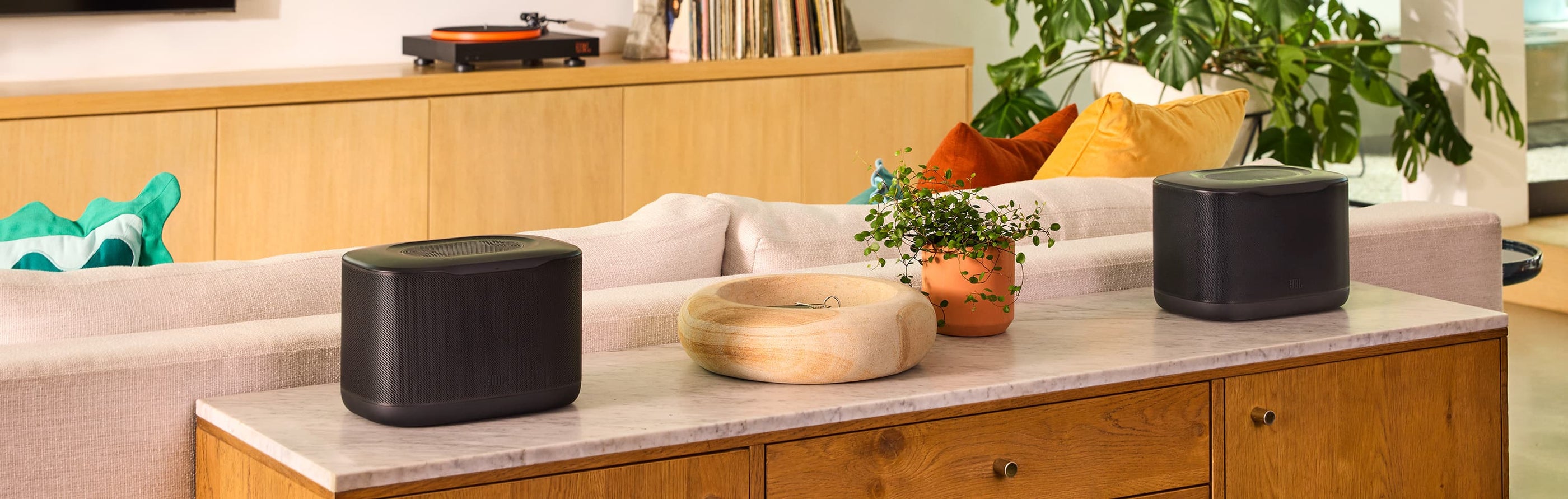 Two JBL Cove X1 wireless speakers displayed on a console table in a modern living room with a JBL turntable in the background.
