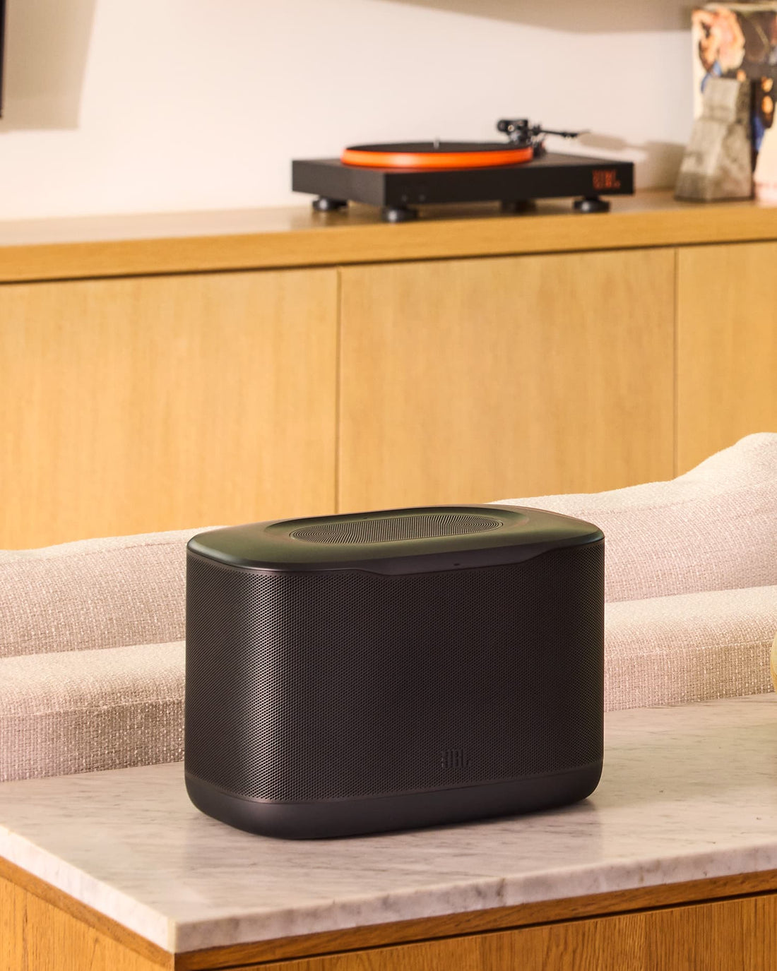 A JBL Cove X1 wireless speaker displayed on a console table in a modern living room with a JBL turntable in the background.
