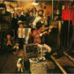 Group of musicians in a basement with instruments and a reel-to-reel tape recorder