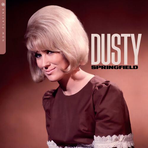 Woman with a blonde bouffant hairstyle wearing a brown dress with lace trim