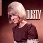 Woman with a blonde bouffant hairstyle wearing a brown dress with lace trim