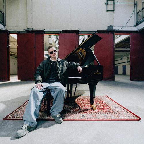 Man in sunglasses and leather jacket seated beside a black grand piano on a patterned rug
