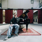 Man in sunglasses and leather jacket seated beside a black grand piano on a patterned rug