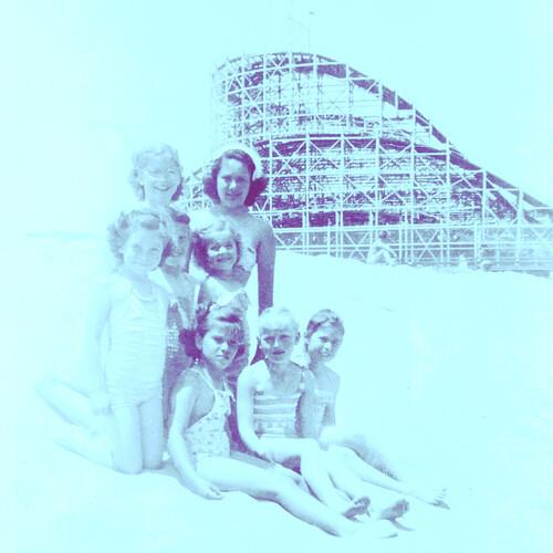 Vintage-style photograph of a group of women in swimsuits posing on a beach with a wooden roller coa