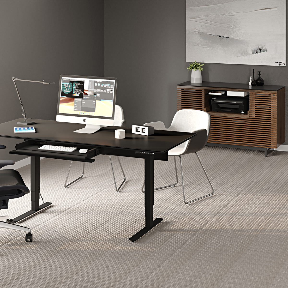 A modern adjustable height standing desk in black with a built-in keyboard tray, styled in a minimalist office setting with a computer, white chairs, and a wooden credenza.
