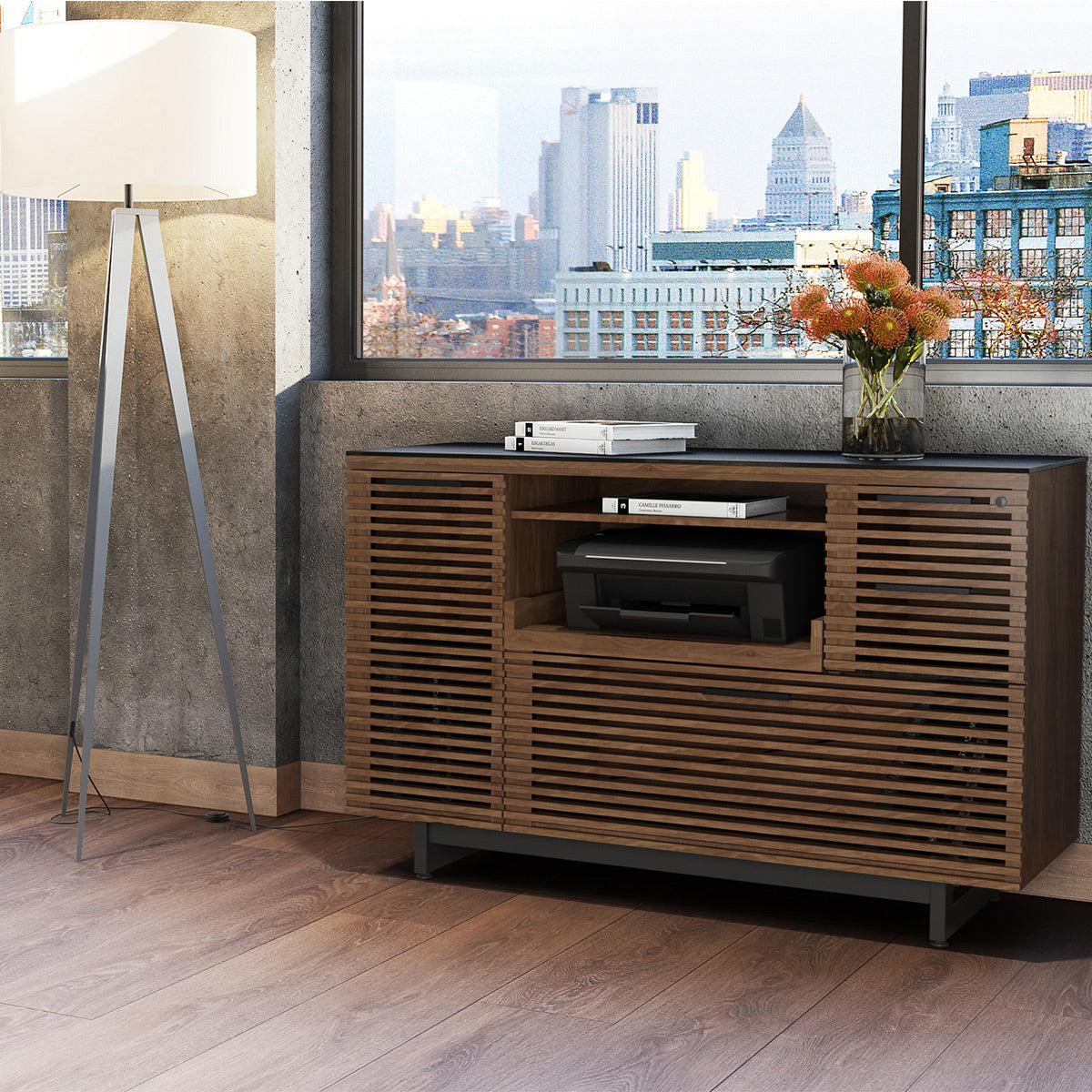 A modern wooden media console cabinet with a dark top, set in a contemporary room with a concrete wall, city skyline window view, a floor lamp, and decorative flowers.